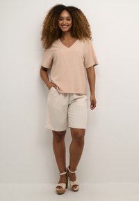 Ljusrosa kortärmad V-ringad topp i kombination med beige knälånga shorts, båda i mjukt tyg. Modellen bär beige kilklackar.
