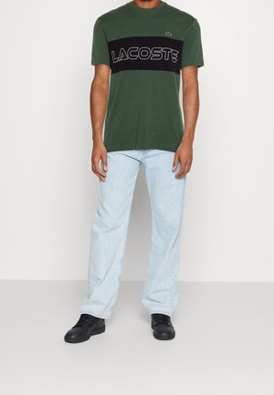T-shirt di cotone verde con una striscia orizzontale nera sulla quale è scritto "LACOSTE" in bianco. Indossato con jeans azzurri chiari e scarpe nere.