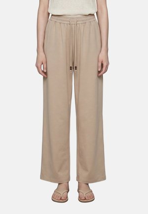 Person trägt beige, weit geschnittene Hosen mit Kordelzug in der Taille und beige Flip-Flops, steht vor einem einfarbigen hellen Hintergrund.