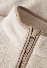Veste en polaire blanc cassé avec fermeture éclair beige et bordure. Présente un tissu doux et texturé avec un col haut.