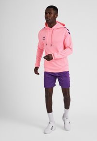 Felpa rosa brillante con un logo, abbinata a pantaloni corti viola. L'outfit include scarpe bianche e calze bianche a vista. Sfondo semplice.