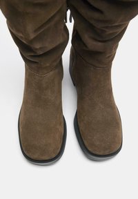 Stivali alla caviglia in suede marrone con un design strutturato, suole nere piatte e dettagli arricciati nella parte superiore per un tocco di texture.