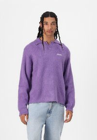 Pulover tricotat violet, pufoasă, cu guler și mâneci lungi, având broderie discretă a logo-ului pe piept.