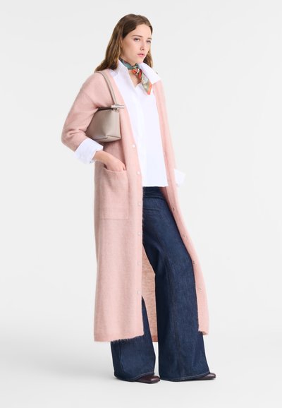 Long pink cardigan made of soft material with button closures and pockets, layered over a white shirt and dark wide-leg jeans.