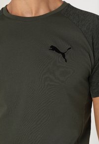 Man i olivgrön och mörkgrå pikétröja med kort ärm från Puma, med svart Puma-logotyp på vänster bröst.