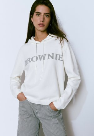 Sudadera blanca con "BROWNIE" en texto gris, capucha con cordones, textura suave. Combinada con pantalones a rayas en gris y blanco, de corte relajado.