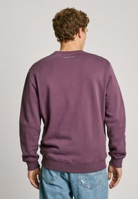 Paarse sweatshirt met ronde hals en lange mouwen. Geribde boorden en zoom. Kleine witte tekst op de achterkant leest "LONDEN W1." Ontworpen voor een ontspannen pasvorm.