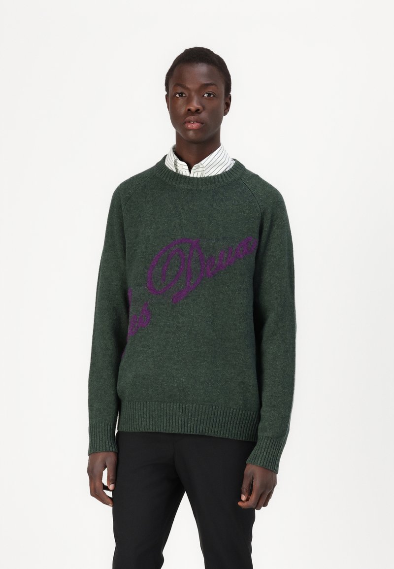Grüner Strickpullover mit gerippten Bündchen und Saum, mit lila Textdesign. Getragen über einem weiß-grün gestreiften Kragenhemd.