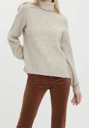 Femme portant un pull à col roulé côtelé beige et un pantalon marron ajusté, posant avec une main derrière le dos.