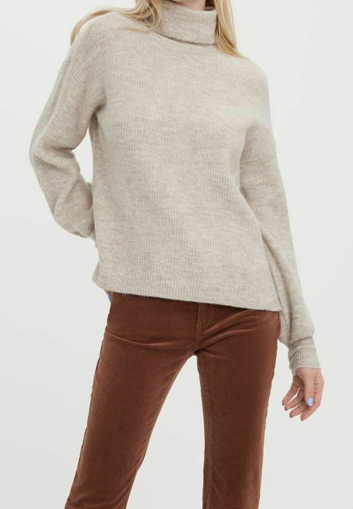 Femme portant un pull à col roulé côtelé beige et un pantalon marron ajusté, posant avec une main derrière le dos.