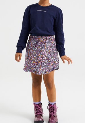 Enfant portant un haut à manches longues bleu marine avec le texte « la gentillesse compte », une jupe fleurie, des chaussettes violettes et des bottines roses brillantes à lacets, debout.
