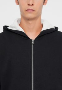 Sweatshirt preto com fecho zip e capuz forrado a branco, apresentando uma textura suave e um fecho de correr prateado. Design simples com um corte moderno.