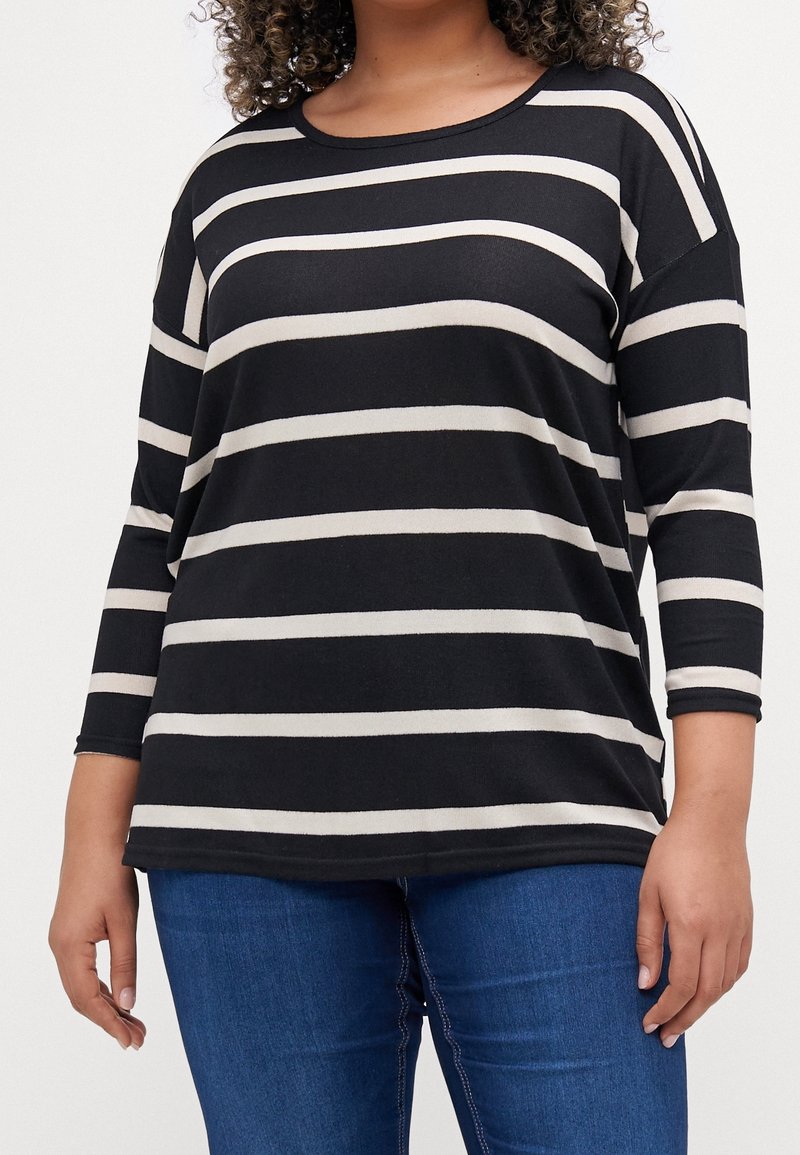 Femme portant un t-shirt à manches longues à rayures horizontales noires et blanches avec un jean bleu, debout devant un fond uni.