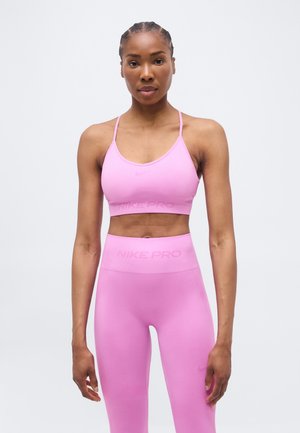 Donna con capelli intrecciati che indossa un reggiseno sportivo rosa Nike Pro e leggings a vita alta, in piedi davanti a uno sfondo grigio chiaro uniforme.