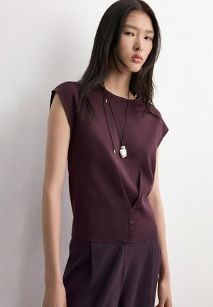 Młoda kobieta o długich ciemnych włosach ubrana w bezrękawnik w kolorze śliwkowym z guzikami oraz dopasowane spodnie, stojąca na tle białej ściany.