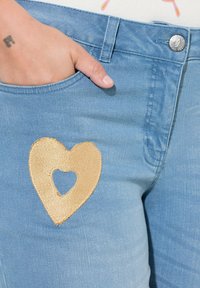 Jeans de mezclilla azul claro con un gran parche de corazón bordado en dorado que incluye una forma de corazón recortada. Se puede ver el bolsillo con un botón metálico.