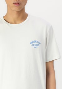 Svetlo šedé bavlnené tričko s modrou výšivkou loga "WRANGLER JEANS 1947" na ľavej hrudi. Klasický okrúhly výstrih a krátke rukávy.