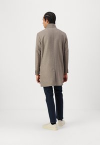 Lys brun ullfrakk med høy krage, rette linjer og sidespalter, kombinert med mørke jeans og hvite sneakers. Teksturert stoffdetalj synlig.
