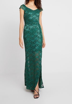 Femme portant une robe longue en dentelle vert émeraude avec manches courtes et fente à l'avant, associée à des talons noirs à brides.