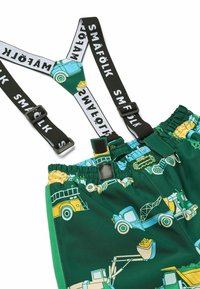 Pantaloni corti verdi con motivi di camion colorati, dotati di bretelle nere con il testo "SMÅFÖLK", vita elastica e una texture liscia.