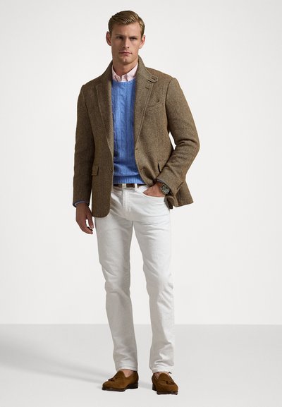 Muškarac stoji s rukom u džepu, odjeven u smeđi sako u riblju kost, plavi džemper od pletene vune, bijele hlače i smeđe cipele s resicama.