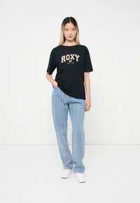 Naine, kellel on seljas must "ROXY" t-särk, heledate siniste triibuliste teksade ja valgete sportkingadega, seisab lihtsalt valge tausta ees.