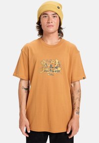 Bruine katoenen t-shirt met een kleurrafloriemotief. Ontspannen pasvorm met korte mouwen. De drager heeft een gele gebreide muts en zichtbare tatoeages.