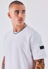 Joven con cabello corto que lleva una camiseta blanca con un detalle de bolsillo y una cadena, mirando hacia un lado, mostrando su brazo tatuado.