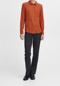 Chemise boutonnée à manches longues de couleur orange brûlé avec col et poche poitrine unique, associée à un pantalon noir à jambes droites et des chaussures brillantes.