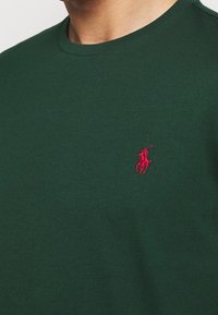 Grön bomullströja med rund halsringning och en liten röd broderad logotyp på vänster bröst. Slät textur med en figurnära design.