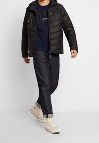 Svart pufferjacka med huva, med en dragkedja fram, quiltat mönster, bärs över en marinblå crew-neck sweatshirts och mörk denimjeans.