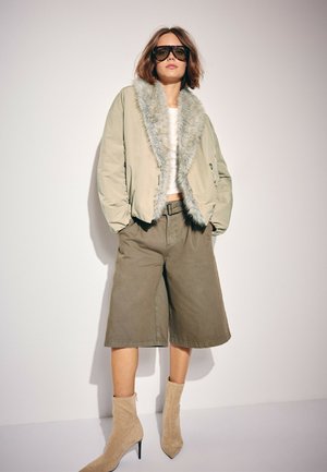 Femme portant une veste beige oversize avec bordure en fourrure, un haut blanc, un short marron large à hauteur des genoux, des bottes beige à talons, et de grandes lunettes de soleil foncées.
