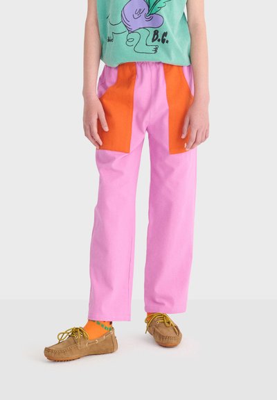 Enfant portant un pantalon rose avec de grandes poches orange, un t-shirt vert à motif, des chaussettes orange et des mocassins marron, debout sur un sol blanc.