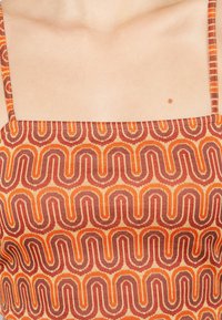 Haut d'un vêtement présentant un motif ondulé en orange et bordeaux sur un fond crème. Les bretelles réglables sont visibles.