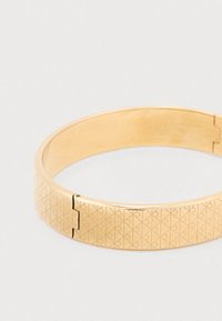 Bracelet en métal doré avec une finition lisse et des motifs géométriques gravés, doté d'une fermeture à charnière pour un enfilage facile.