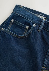 Tmavomodré rifle z denimu s prednými vreckami, nitmi, slučkou na opasok a viditeľnou štítkou "Next Denim Supply" na páse.