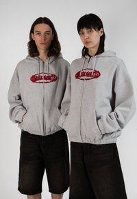 Sweatshirts zippés gris avec logo rouge brodé, présentant une coupe décontractée et des poignets élastiques, associés à un short en denim noir.