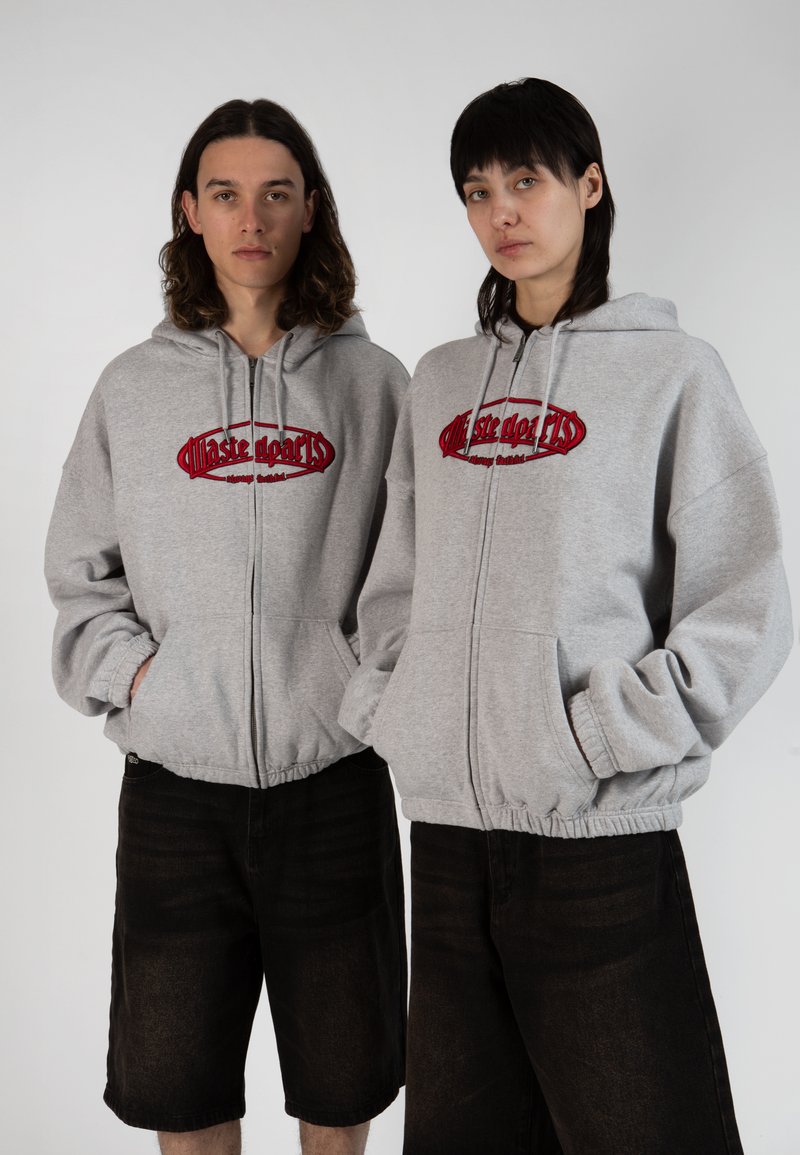 Sweatshirts zippés gris avec logo rouge brodé, présentant une coupe décontractée et des poignets élastiques, associés à un short en denim noir.