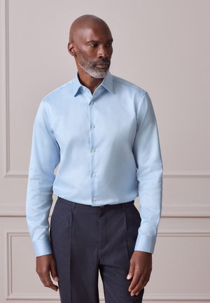 Hombre con barba gris vestido con camisa azul clara y pantalones oscuros, de pie contra una pared beige, mirando hacia un lado.