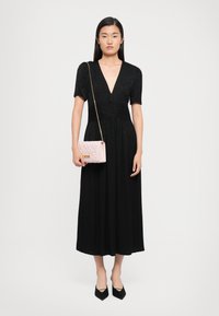 Donna che indossa un vestito midi nero con maniche corte, scollatura a V profonda, scarpe nere a punta e tiene in mano una borsa a tracolla trapuntata rosa chiaro con catena dorata.