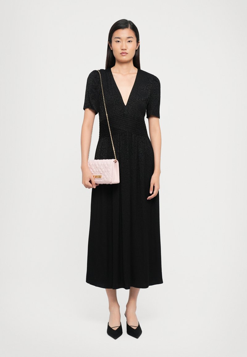 Donna che indossa un vestito midi nero con maniche corte, scollatura a V profonda, scarpe nere a punta e tiene in mano una borsa a tracolla trapuntata rosa chiaro con catena dorata.