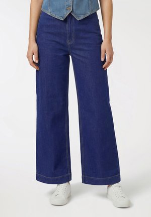 Osoba ubrana w ciemnoniebieskie dżinsy o wysokim stanie i szerokich nogawkach, białe trampki oraz krótką, niebieską kamizelkę jeansową, stojąca z rozluźnionymi ramionami.