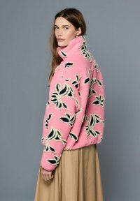 Roza flis jakna z visokim ovratnikom, okrašena s črno in kremasto cvetlično vezenino. Model nosi dolgo bež krilo, pogled iz strani.