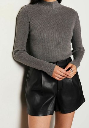 Femme portant un pull à manches longues gris côtelé avec des boutons sur l'épaule et un short en cuir noir taille haute, les mains jointes.