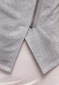 Světle šedý textilní oděv s uzávěrem na zip a texturovaným geometrickým vzorem, s vrstveným designem a viditelnou měkkou vnitřní látkou.