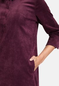 Frau in einem lila Cordkleid mit langen Ärmeln und Knöpfen, die Hand in die seitliche Tasche gesteckt, vor einem schlichten Hintergrund.