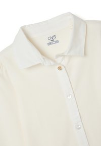 Cream-colored shirt with a collared neck, button-down front, and short sleeves. Features a smooth texture and subtle logo branding inside the collar.