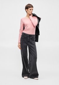 Ružová blúzka s dlhým rukávom skombinovaná s tmavošedými džínsami so širokými nohavicami, ktoré majú predné vrecká a viditeľné švy. Modelka drží čiernu bundu.