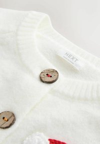 Gros plan d'un cardigan blanc duveteux avec des boutons en bois et une étiquette de vêtement visible indiquant « NEXT Made in China. »