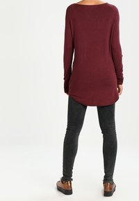 Maglietta bordeaux a maniche lunghe con scollo rotondo, caratterizzata da un orlo curvo. Abbinata a jeans skinny grigi scuri e scarpe marroni.